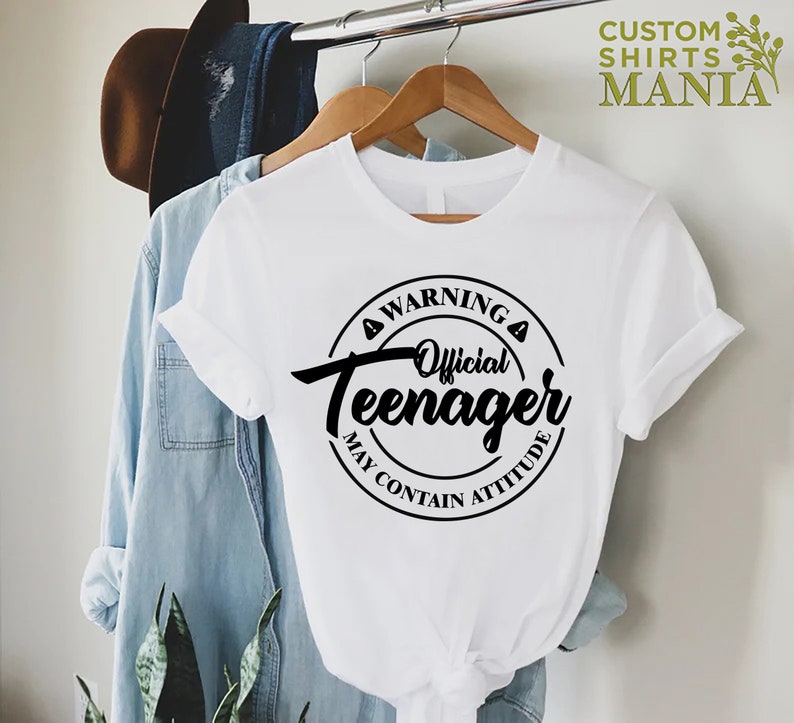May include: White t-shirt with a black circle graphic that says "Warning Official Teenager May Contain Attitude".