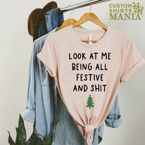May include: A pink t-shirt with the text "Look at me being all festive and shit" and a small green Christmas tree with lights.