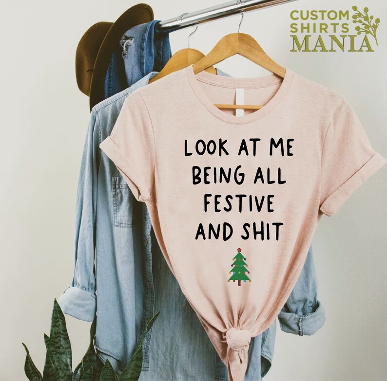 May include: A pink t-shirt with the text "Look at me being all festive and shit" and a small green Christmas tree with lights.