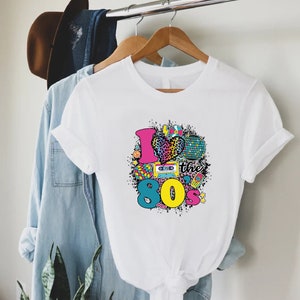 May include: White t-shirt with a graphic print of a heart with a leopard print, a cassette tape, a disco ball, and the text "I love the 80s".