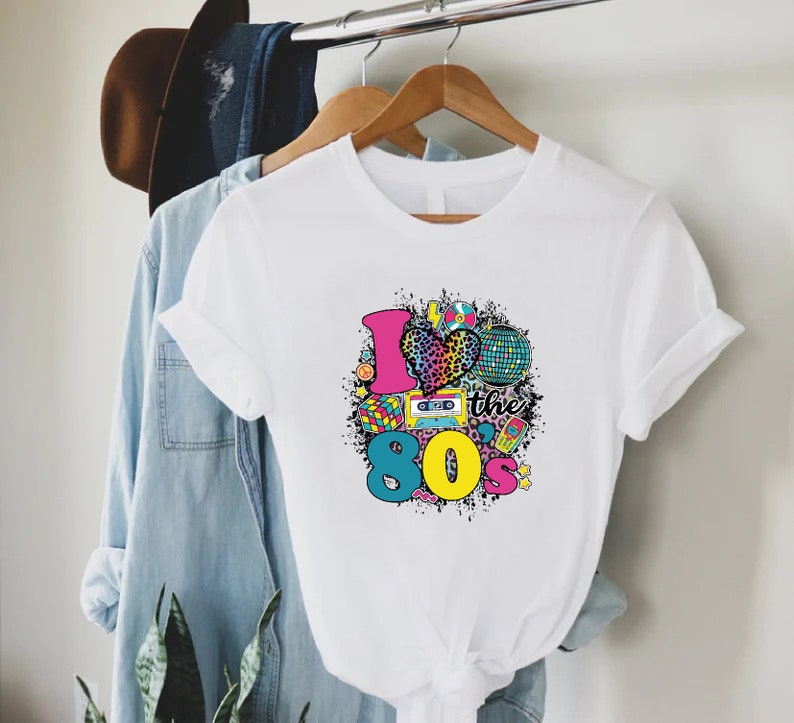 May include: White t-shirt with a graphic print of a heart with a leopard print, a cassette tape, a disco ball, and the text "I love the 80s".