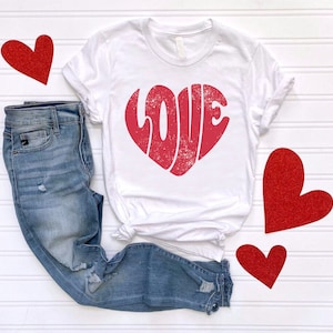 May include: White t-shirt with a red heart-shaped graphic that says "LOVE" in a vintage style. The shirt is paired with blue jeans and red glitter hearts.