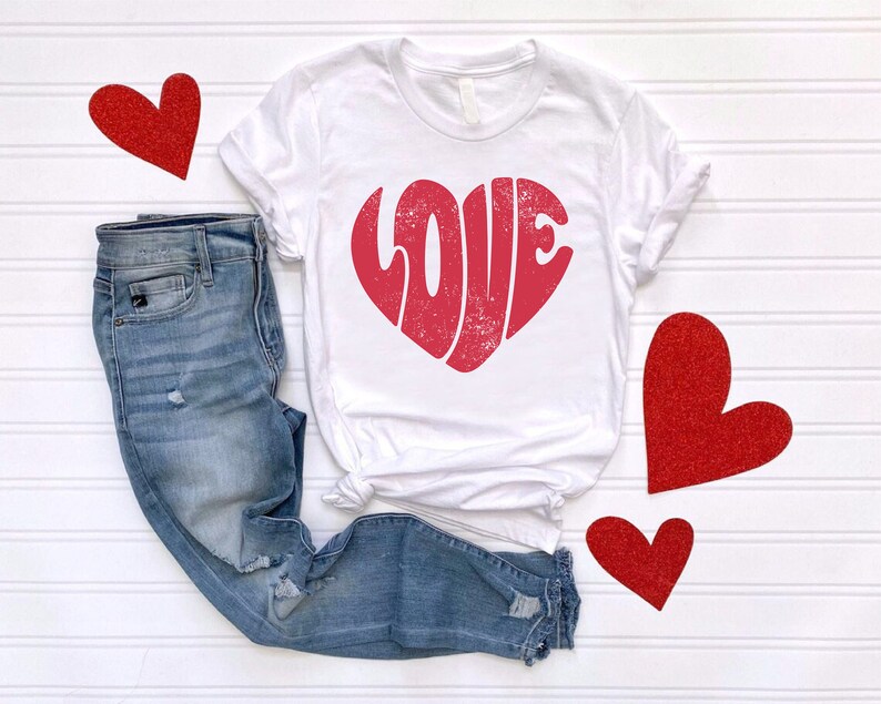 May include: White t-shirt with a red heart-shaped graphic that says "LOVE" in a vintage style. The shirt is paired with blue jeans and red glitter hearts.