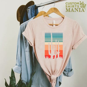 May include: A light pink t-shirt with a retro-style graphic of a cactus silhouette against a colorful striped background. The text "CUSTOM SHIRTS MANIA" is visible in the top right corner of the image.