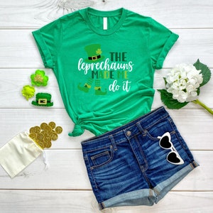 May include: Green t-shirt with a white graphic that says "The Leprechauns Made Me Do It". The graphic also includes a cartoon leprechaun with a green hat and a pot of gold. The shirt is paired with blue denim shorts and white sunglasses.
