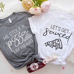Puede incluir: Dos camisetas grises y blancas con un gr&aacute;fico de pizza y el texto "He stole a pizza my heart" y "Let's get sauced".