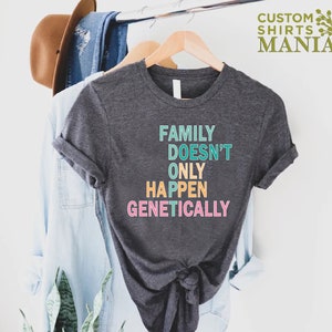 Op de afbeelding: Een donkergrijs T-shirt met de tekst "FAMILY DOESN'T ONLY HAPPEN GENETICALLY" in regenboogkleuren.