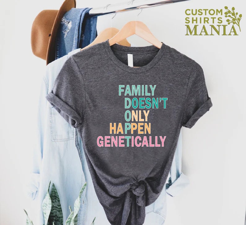 Op de afbeelding: Een donkergrijs T-shirt met de tekst "FAMILY DOESN'T ONLY HAPPEN GENETICALLY" in regenboogkleuren.
