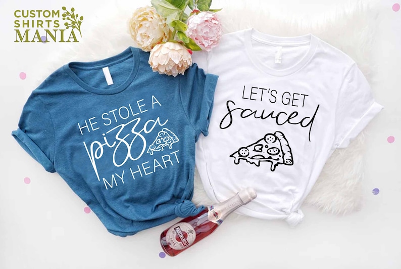 Puede incluir: Dos camisetas con tem&aacute;tica de pizza. Una camiseta es azul turquesa con el texto "Se rob&oacute; una pizza mi coraz&oacute;n" y un peque&ntilde;o gr&aacute;fico de pizza. La otra camiseta es blanca con el texto "Vamos a emborracharnos" y un gr&aacute;fico de pizza m&aacute;s grande.