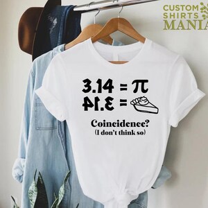 K&ouml;nnte beinhalten: Wei&szlig;es T-Shirt mit schwarzem Text, der "3.14 = &pi; &pi;.&epsilon; =" lautet, mit einer Tortendarstellung. Der Text darunter lautet "Zufall? (Ich glaube nicht)"