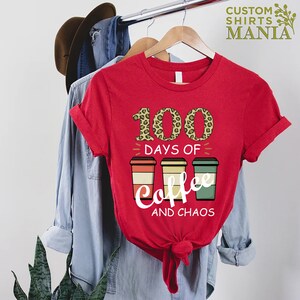 Pu&ograve; includere: Maglietta rossa con il testo "100 giorni di caff&egrave; e caos" in bianco. Il numero 100 &egrave; in stampa leopardata. Ci sono tre tazze di caff&egrave; con disegni retr&ograve;.