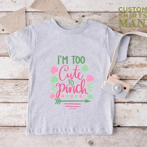 May include: A gray t-shirt with the text "I'm Too Cute To Pinch" in pink and green lettering. The design includes shamrocks, an arrow, and stripes.