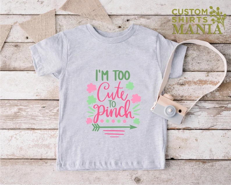May include: A gray t-shirt with the text "I'm Too Cute To Pinch" in pink and green lettering. The design includes shamrocks, an arrow, and stripes.
