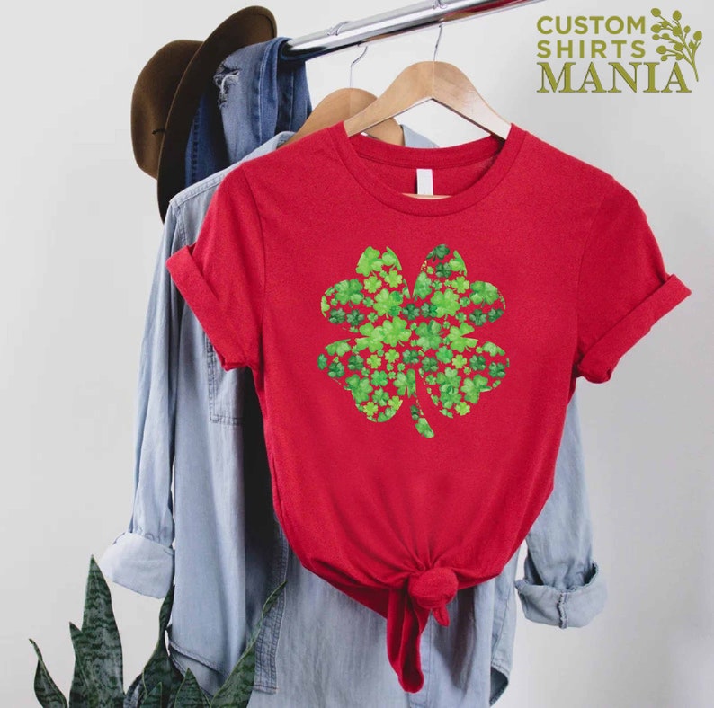 May include: Red t-shirt with a green four-leaf clover design made of smaller four-leaf clovers. The shirt is hanging on a hanger.