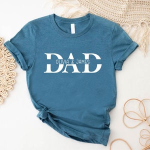 Pu&ograve; includere: Una maglietta blu turchese con la parola "DAD" in lettere bianche, con una linea sotto la parola e il testo "OLIVIA & JAMES" sotto.