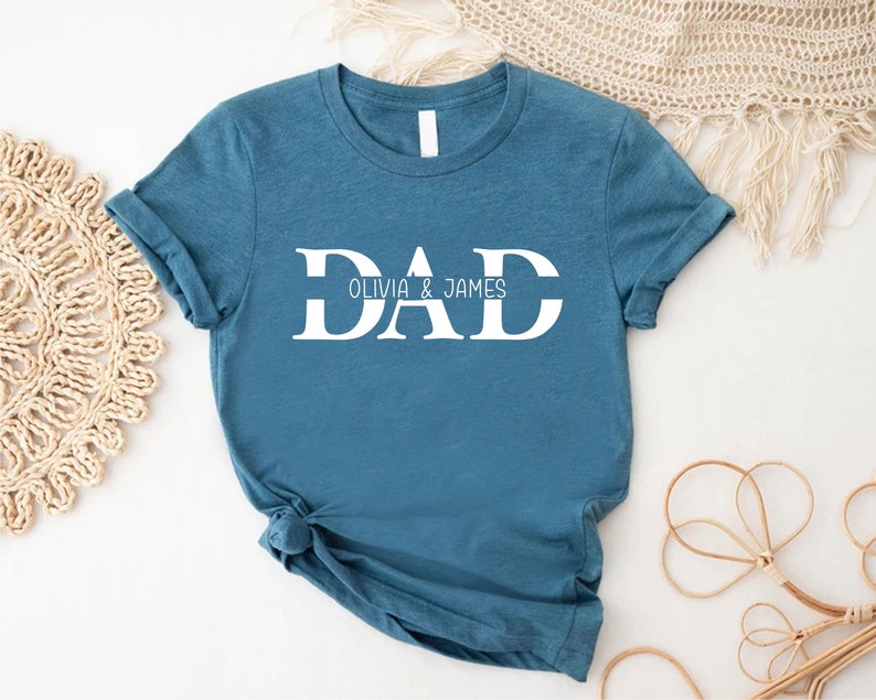 Pu&ograve; includere: Una maglietta blu turchese con la parola "DAD" in lettere bianche, con una linea sotto la parola e il testo "OLIVIA & JAMES" sotto.