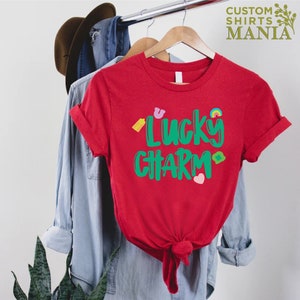 May include: Red t-shirt with a green graphic that says "Lucky Charm" and features small colourful icons.
