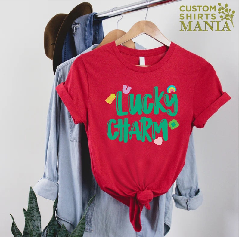 May include: Red t-shirt with a green graphic that says "Lucky Charm" and features small colourful icons.