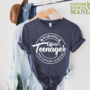 May include: A navy blue t-shirt with a white circle graphic that says "Warning Official Teenager May Contain Attitude".