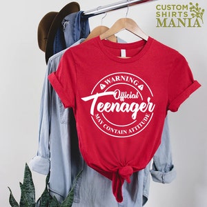 May include: Red t-shirt with a white circle graphic that says "Warning Official Teenager May Contain Attitude".