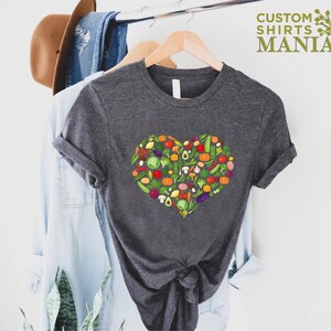 May include: A dark gray t-shirt featuring a heart-shaped design composed of various colorful vegetables. The shirt is hanging on a wooden hanger. The text "CUSTOM SHIRTS MANIA" is visible in the upper right corner.
