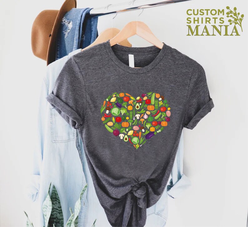 May include: A dark gray t-shirt featuring a heart-shaped design composed of various colorful vegetables. The shirt is hanging on a wooden hanger. The text "CUSTOM SHIRTS MANIA" is visible in the upper right corner.