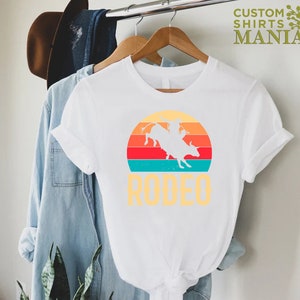 May include: White t-shirt with a vintage-style graphic of a cowboy riding a bull in front of a sunset. The text "RODEO" is printed below the graphic.