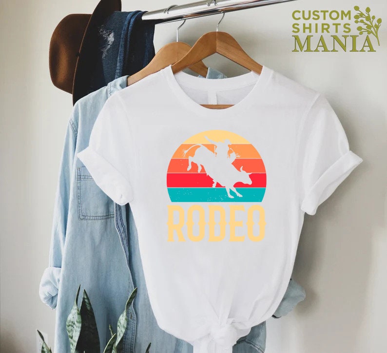 May include: White t-shirt with a vintage-style graphic of a cowboy riding a bull in front of a sunset. The text "RODEO" is printed below the graphic.