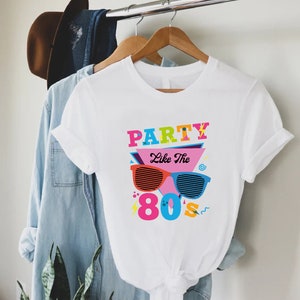 K&ouml;nnte beinhalten: Wei&szlig;es T-Shirt mit einem bunten Grafikdesign, auf dem "Party Like The 80's" mit Sonnenbrille und anderen Designs im 80er-Jahre-Stil steht.