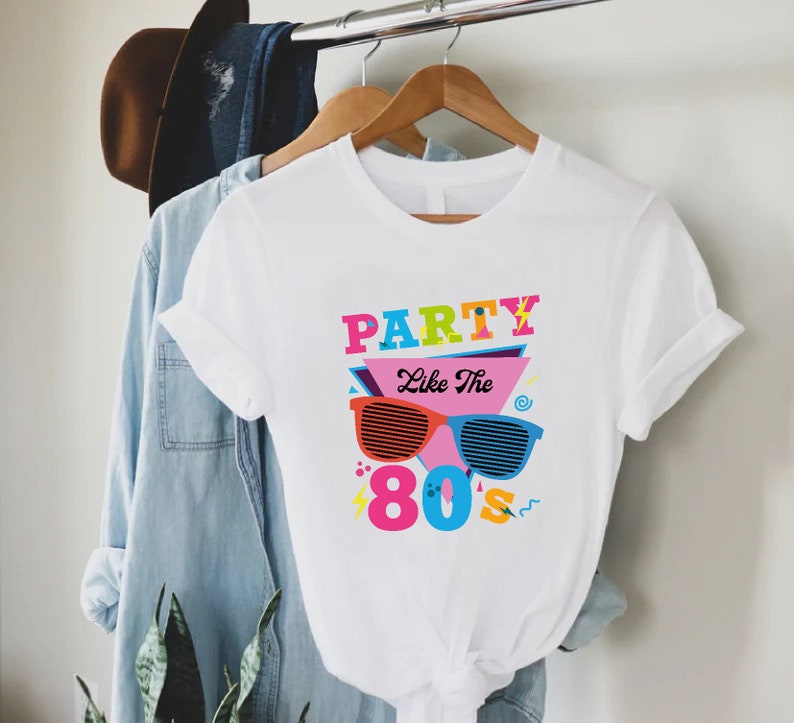 K&ouml;nnte beinhalten: Wei&szlig;es T-Shirt mit einem bunten Grafikdesign, auf dem "Party Like The 80's" mit Sonnenbrille und anderen Designs im 80er-Jahre-Stil steht.