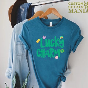 May include: A teal blue t-shirt with the text "Lucky Charm" in green letters. The shirt has a variety of small, colourful graphics, including a rainbow, a shamrock, a horseshoe, and a heart.