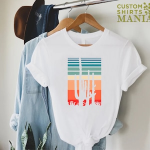 May include: White t-shirt with a retro-style graphic of three cacti in front of a sunset-colored striped background.