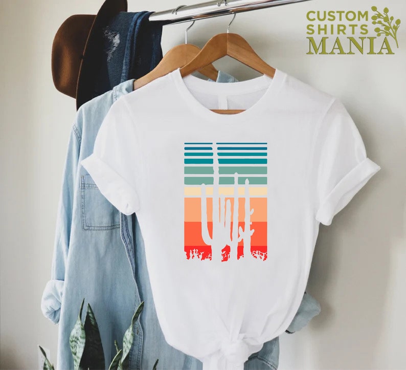 May include: White t-shirt with a retro-style graphic of three cacti in front of a sunset-colored striped background.