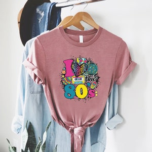 May include: A pink t-shirt with a graphic design that says "I love the 80's" with a heart, a cassette tape, a Rubik's cube, a disco ball, and other 80's themed icons.