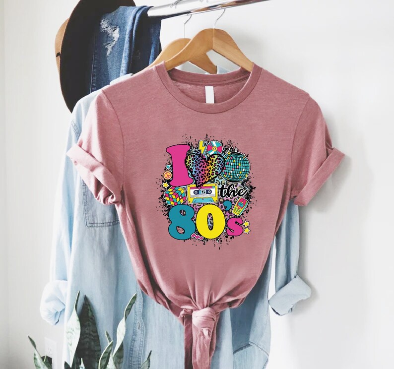 May include: A pink t-shirt with a graphic design that says "I love the 80's" with a heart, a cassette tape, a Rubik's cube, a disco ball, and other 80's themed icons.