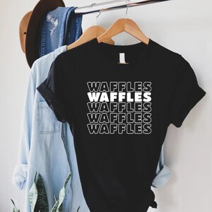 Może przedstawiać: Czarny T-shirt z napisem "Waffles" powt&oacute;rzonym pięć razy na biało, ułożonym pionowo. Tekst jest w pogrubionym, retro kroju.