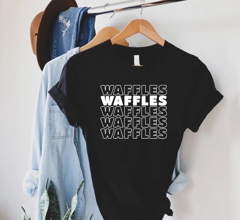 Może przedstawiać: Czarny T-shirt z napisem "Waffles" powt&oacute;rzonym pięć razy na biało, ułożonym pionowo. Tekst jest w pogrubionym, retro kroju.