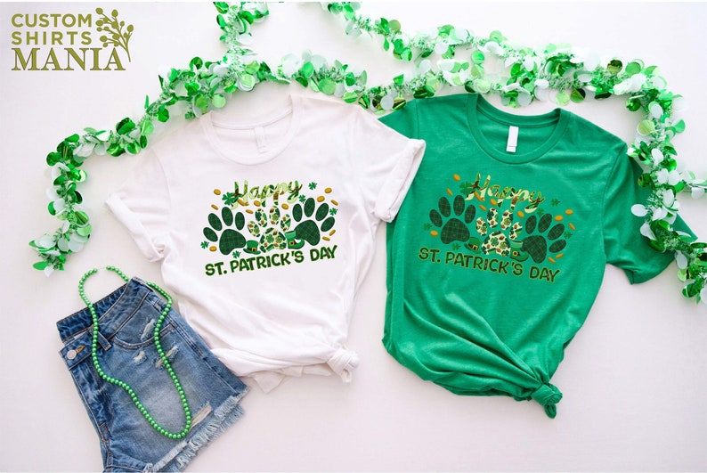 May include: Two t-shirts with a green shamrock and paw print design. The white t-shirt says "Happy St. Patrick's Day" and the green t-shirt says "Happy St. Patrick's Day".