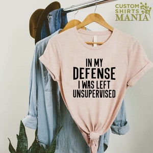 May include: A pink t-shirt with the text "In my defense I was left unsupervised" printed in black.