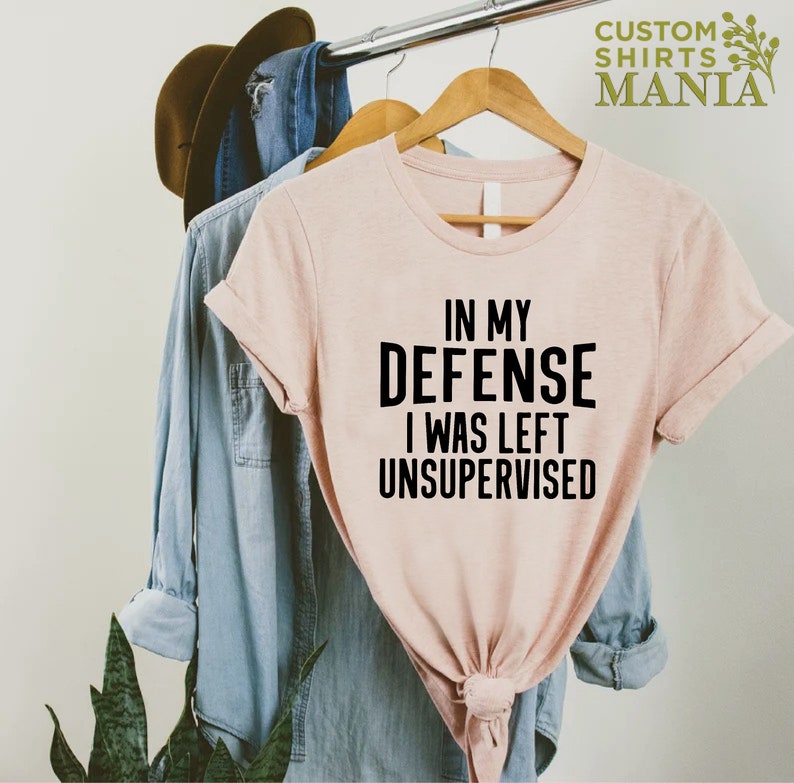 May include: A pink t-shirt with the text "In my defense I was left unsupervised" printed in black.