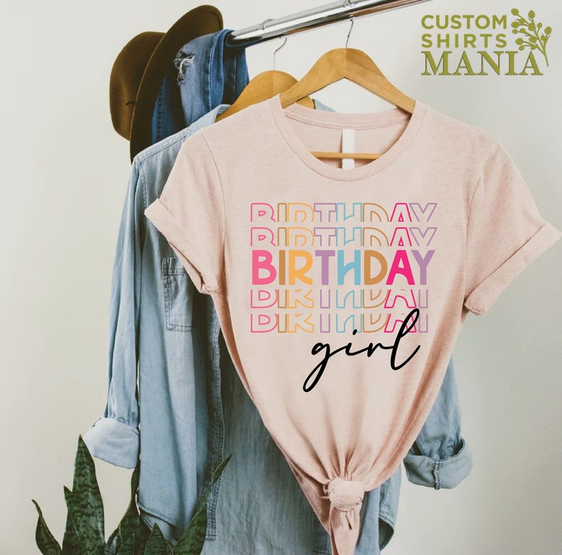 May include: A pink t-shirt with the text "Birthday girl" in rainbow colours. The text is stacked and the word "Birthday" is repeated multiple times.