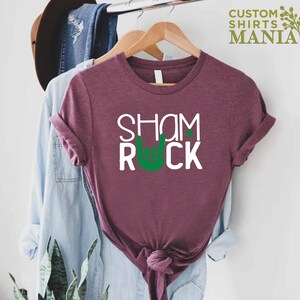 Peut inclure: Un t-shirt marron avec le texte "Shamrock" et un symbole de main de rock and roll vert avec un tr&egrave;fle sur l'index.