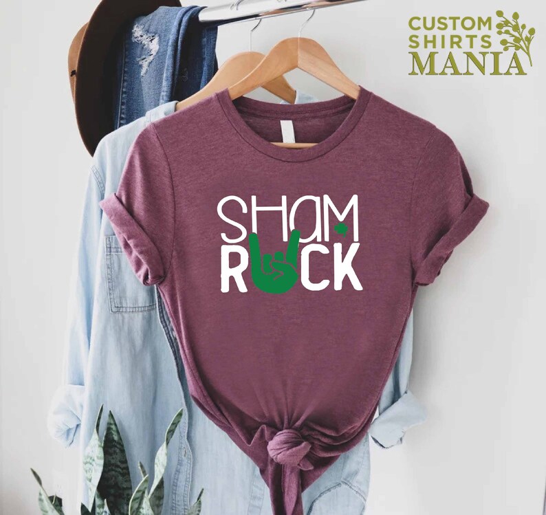 Peut inclure: Un t-shirt marron avec le texte "Shamrock" et un symbole de main de rock and roll vert avec un tr&egrave;fle sur l'index.