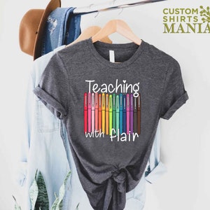 May include: A dark gray t-shirt with the text "Teaching with flair" and a row of colorful markers.