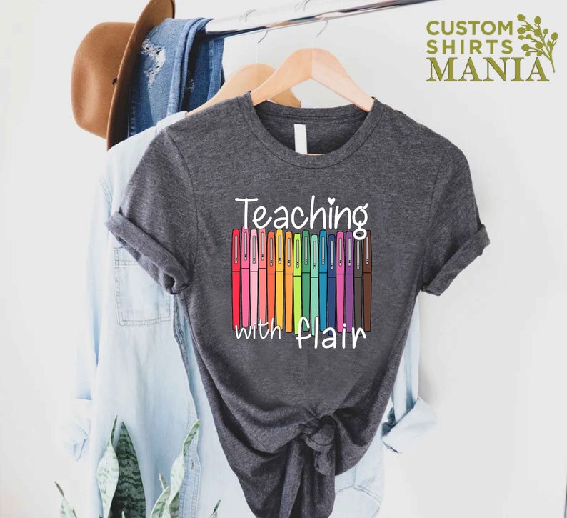 May include: A dark gray t-shirt with the text "Teaching with flair" and a row of colorful markers.