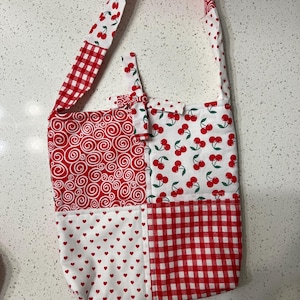 May include: A red and white patchwork tote bag with a cherry print, gingham pattern, and red swirl design. The bag has a long strap and a small pocket on the front.