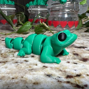 May include: A green segmented frog toy with black eyes. The frog is lying on a light brown surface.