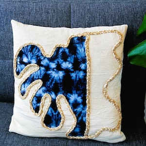Op de afbeelding: Een wit kussen met een blauw en wit tie-dye patroon en een gouden touwrand. Het kussen ligt op een blauwe bank.