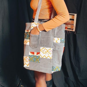 Peut inclure: Une femme portant une chemise à manches longues orange brûlée et un pantalon noir tient un sac cabas gris avec un motif patchwork. Le sac est en jean et présente une variété de motifs géométriques colorés.