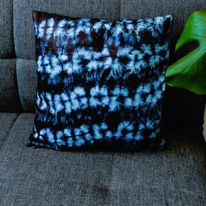 Op de afbeelding: Een blauw en zwart tie-dye kussen met een witte achtergrond. Het kussen ligt op een grijze bank met een groene plant op de achtergrond.